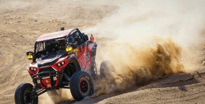 A racecar jeep is being driven across a dirt plain, making a turn ot the right. Dust is swept up in the turn.