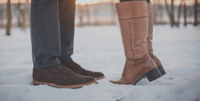 A view from the ground showing the lower legs of a couple facing each other and standing in the snow. One is wearing gray pants and brown shoes, and the other is wearing tall tan boots. The second person is standing on their heels.