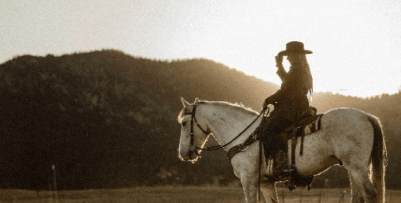 A woman wearing cowboy apparel sitting on a white horse. She is tilting her hat. The background shows a mountain with the sunset resting against it.