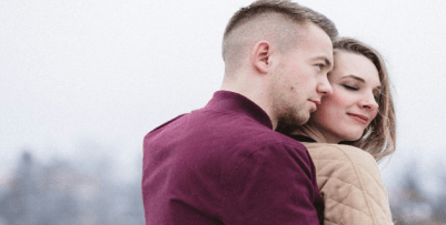 A man in a red shirt romantically holding a woman in a tan sweater from behind her. The man looks forward, and the woman is smiling with her eyes closed.