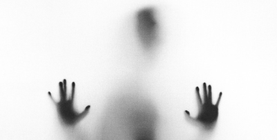 A humanoid figure hidden behind fogged glass. The vague shape of their head and torso is visible. Their hands are pressed up against the glass.