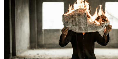 A man holding a newspaper that is on fire. He is holding the newspaper up to cover his face.