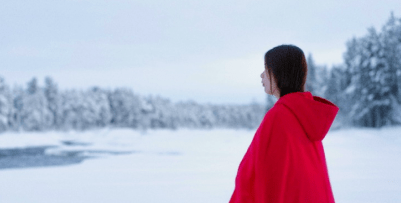 A woman is standing in a forest covered in snow. She is wearing a red parka that contrasts against the white of the snow.