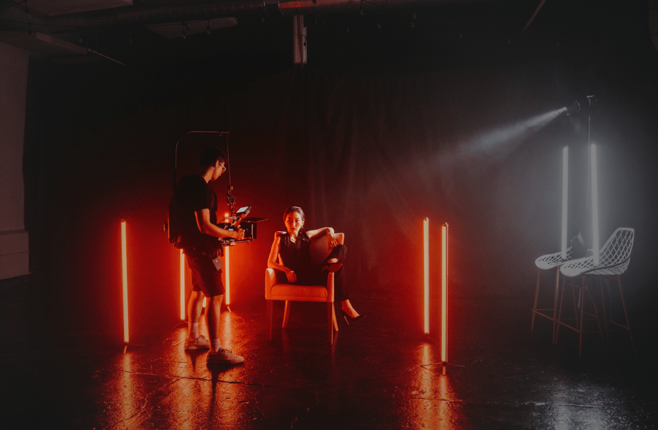 A woman sitting sideways on an armchair with her legs over the armrest, posing casually. A man holds a camera pointed at her. The room is lit up in red and white light, giving a dramatic atmosphere.